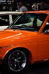 An orange Datsun car is prominently displayed, featuring sleek lines and polished chrome wheels. In the background, a white car is partially visible, along with a few people standing and conversing near a display area, possibly at a car show or outdoor event.