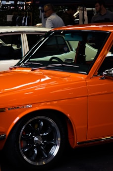 An orange Datsun car is prominently displayed, featuring sleek lines and polished chrome wheels. In the background, a white car is partially visible, along with a few people standing and conversing near a display area, possibly at a car show or outdoor event.