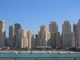 A row of impressive skyscrapers lines the waterfront, with modern architectural designs. Below the towering buildings, a sandy beach is visible, populated by people engaging in leisurely activities. Several yachts and boats are anchored in the turquoise water, enhancing the luxurious atmosphere.