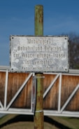 A metallic sign is mounted on a rusty green pole, displaying instructions in German that unauthorized access is prohibited. The background features a blurred view of a wooden fence and a clear blue sky.