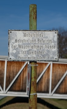 A metallic sign is mounted on a rusty green pole, displaying instructions in German that unauthorized access is prohibited. The background features a blurred view of a wooden fence and a clear blue sky.