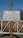 A metallic sign is mounted on a rusty green pole, displaying instructions in German that unauthorized access is prohibited. The background features a blurred view of a wooden fence and a clear blue sky.