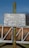 A metallic sign is mounted on a rusty green pole, displaying instructions in German that unauthorized access is prohibited. The background features a blurred view of a wooden fence and a clear blue sky.