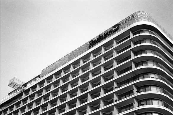 A modern hotel building with a curvilinear architectural design featuring multiple floors of balcony spaces. The structure emphasizes horizontal lines and geometric patterns, creating a sleek, uniform appearance. The photograph is captured in black and white, highlighting the contrast and architectural details.