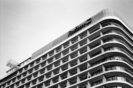 A modern hotel building with a curvilinear architectural design featuring multiple floors of balcony spaces. The structure emphasizes horizontal lines and geometric patterns, creating a sleek, uniform appearance. The photograph is captured in black and white, highlighting the contrast and architectural details.