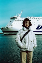 A person wearing a white winter jacket stands in front of a large ferry named 'Polonia' on a calm body of water. The person appears to be smiling, with hands in pockets, and the background features a clear blue sky.