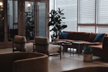 A modern lounge area featuring mid-century style furniture, including a large brown sectional sofa with blue cushions, beige armchairs, and wooden coffee tables. The space is adorned with tall green plants and illuminated by natural light filtering through venetian blinds.