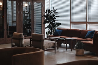 A modern lounge area featuring mid-century style furniture, including a large brown sectional sofa with blue cushions, beige armchairs, and wooden coffee tables. The space is adorned with tall green plants and illuminated by natural light filtering through venetian blinds.