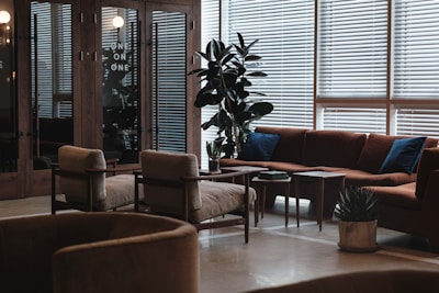 A modern lounge area featuring mid-century style furniture, including a large brown sectional sofa with blue cushions, beige armchairs, and wooden coffee tables. The space is adorned with tall green plants and illuminated by natural light filtering through venetian blinds.