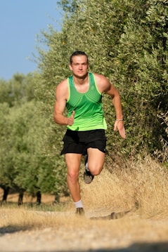 A man is running towards the camera on a dirt path surrounded by trees. He is wearing a bright green sleeveless top and black shorts. The backdrop of lush green trees and a clear blue sky suggests a sunny day, ideal for outdoor activities.