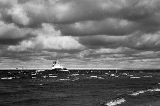 A detailed black and white photograph of a World War II battleship cutting through choppy seas under a cloudy sky.