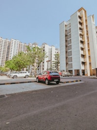 A parking lot in front of high-rise residential buildings with several parked cars including a prominent red car. The sky is clear and blue, with green trees adding a touch of nature to the urban setting.
