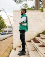 A person with braided hair stands confidently on outdoor steps, wearing a turquoise vest, black cargo pants, and black boots. A transparent bag with a black strap is worn across the shoulder. The background features a light stone wall, greenery, and a blurred view of a roadway with cars and trees.