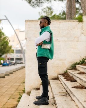 A person with braided hair stands confidently on outdoor steps, wearing a turquoise vest, black cargo pants, and black boots. A transparent bag with a black strap is worn across the shoulder. The background features a light stone wall, greenery, and a blurred view of a roadway with cars and trees.