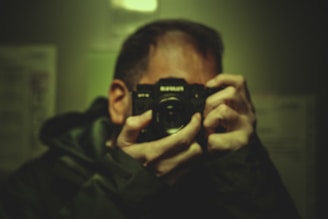 A person holds a camera up to their face, focusing on capturing an image. The scene is dimly lit with a greenish hue, creating a moody atmosphere. The camera is prominently featured in the foreground.