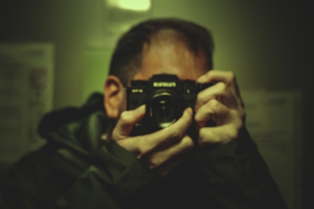 A person holds a camera up to their face, focusing on capturing an image. The scene is dimly lit with a greenish hue, creating a moody atmosphere. The camera is prominently featured in the foreground.