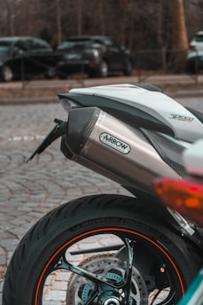 A close-up view of the rear section of a motorcycle with a focus on the exhaust pipe labeled 'Arrow Special Parts'. The motorcycle tire is prominent, showing the Michelin branding on the side. In the background, there are parked cars on a cobblestone surface, with trees lining the scene.