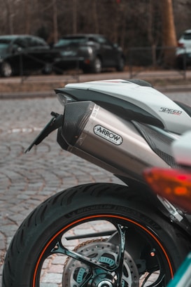 A close-up view of the rear section of a motorcycle with a focus on the exhaust pipe labeled 'Arrow Special Parts'. The motorcycle tire is prominent, showing the Michelin branding on the side. In the background, there are parked cars on a cobblestone surface, with trees lining the scene.