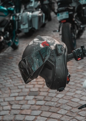 A motorcycle helmet is seen resting on the handlebars of a parked motorcycle. The helmet has a dark, intricate design with a red rose graphic. It has a tinted visor, reflecting the surrounding environment. Cobblestone pavement and multiple motorcycles are visible in the background.