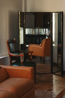 A room with elegant vintage furniture, including a brown leather sofa and green upholstered chairs with wooden frames. The chairs are positioned in front of a large ornate mirror that reflects the room's interior.