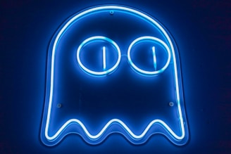 A blue neon sign resembling a ghost figure with two large circular eyes. The glow from the neon tubing creates a bright outline against a dark background.