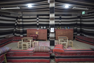 An indoor setup featuring traditional seating arrangements with red and patterned cushions and low wooden tables. The walls are decorated with striped fabric and there are various objects such as pots, kettles, and other containers placed on wooden chests at the back. The ceiling is also adorned with striped fabric and the area is illuminated by lighting fixtures.