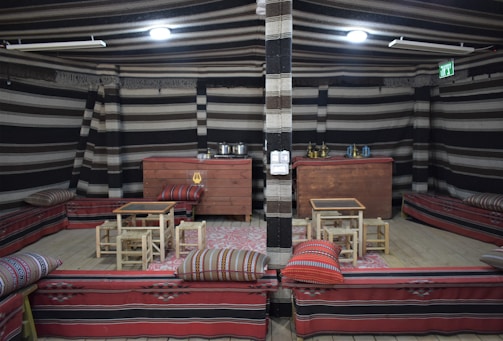 An indoor setup featuring traditional seating arrangements with red and patterned cushions and low wooden tables. The walls are decorated with striped fabric and there are various objects such as pots, kettles, and other containers placed on wooden chests at the back. The ceiling is also adorned with striped fabric and the area is illuminated by lighting fixtures.