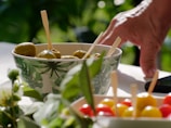 A hand reaches towards a small bowl filled with green olives, each skewered with a wooden stick. The bowl has a green leaf pattern and is situated on a table. In the foreground, a cluster of small, colorful tomatoes, and some green plant leaves are visible, with a soft focus. The lighting is bright and natural, suggesting an outdoor setting.