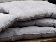 A stack of folded grey cushions placed on a wooden surface. The cushions have a textured fabric with visible seams and appear thick and comfortable.