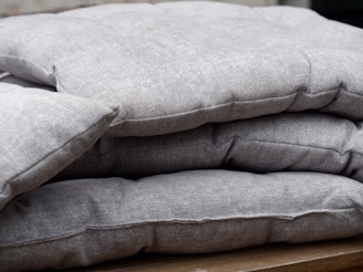 A stack of folded grey cushions placed on a wooden surface. The cushions have a textured fabric with visible seams and appear thick and comfortable.