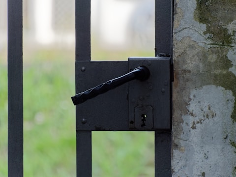 A close-up view of a black metal latch on a gate. The gate is attached to a weathered stone or concrete post, which shows signs of wear and age. Softly blurred green grass can be seen in the background, suggesting an outdoor setting.