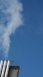 Several industrial chimneys emit white smoke against a clear blue sky. The chimneys are positioned on an industrial building structure, with metal pipes visible.
