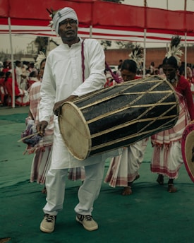 A person in traditional attire is playing a large drum with a group of other performers in the background. They wear white clothing and a turban with a feather decoration. The setting appears to be a cultural or festive event, with a red and white canopy overhead and other participants dressed similarly in white outfits.