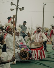 A group of people dressed in traditional attire participate in a cultural event. They are playing musical instruments, including drums, and some are wearing white clothes with red stripes and headgear adorned with feathers. The background features a few people and loudspeakers attached to a utility pole.