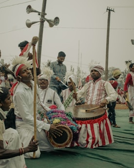 A group of people dressed in traditional attire participate in a cultural event. They are playing musical instruments, including drums, and some are wearing white clothes with red stripes and headgear adorned with feathers. The background features a few people and loudspeakers attached to a utility pole.