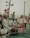 A group of people dressed in traditional attire participate in a cultural event. They are playing musical instruments, including drums, and some are wearing white clothes with red stripes and headgear adorned with feathers. The background features a few people and loudspeakers attached to a utility pole.