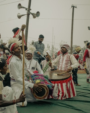 A group of people dressed in traditional attire participate in a cultural event. They are playing musical instruments, including drums, and some are wearing white clothes with red stripes and headgear adorned with feathers. The background features a few people and loudspeakers attached to a utility pole.