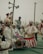 A group of people dressed in traditional attire participate in a cultural event. They are playing musical instruments, including drums, and some are wearing white clothes with red stripes and headgear adorned with feathers. The background features a few people and loudspeakers attached to a utility pole.