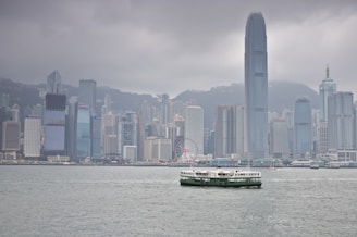 A city skyline featuring a cluster of tall skyscrapers with diverse architectural designs. In the foreground, a ferry is navigating across a broad expanse of water under a cloudy sky.