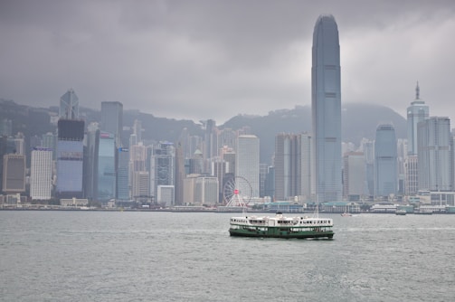 A city skyline featuring a cluster of tall skyscrapers with diverse architectural designs. In the foreground, a ferry is navigating across a broad expanse of water under a cloudy sky.