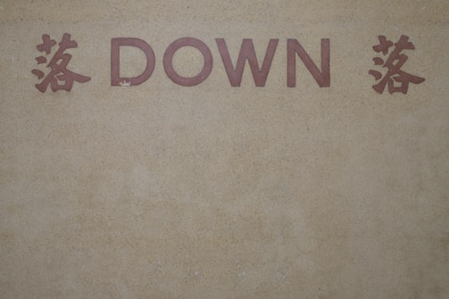 The image features the word 'DOWN' in bold, capitalized, red letters set against a textured, beige background. On either side of the word are red Chinese characters that add an additional element of cultural design.
