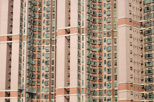 A densely packed facade of a high-rise residential building with numerous windows spaced evenly across multiple floors. The building features a repetitive pattern with light brown and beige exterior colors, contrasted by green-tinted window glass. Air conditioning units are visible below many of the windows.
