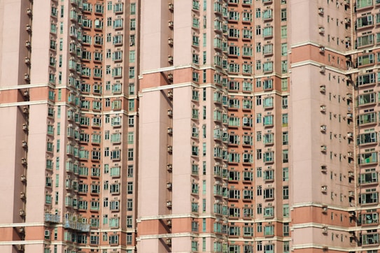 A densely packed facade of a high-rise residential building with numerous windows spaced evenly across multiple floors. The building features a repetitive pattern with light brown and beige exterior colors, contrasted by green-tinted window glass. Air conditioning units are visible below many of the windows.