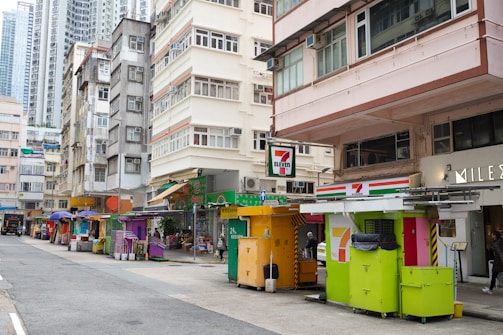A busy urban street scene with multi-story residential buildings. Several colorful kiosks line the sidewalk, offering various goods and services. The prominent 7-Eleven sign is visible, indicating a convenience store nearby. People are walking past, indicating a daily bustling activity. Medium-rise buildings are closely packed, characteristic of a densely populated city area.