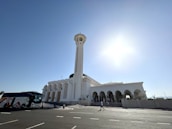 A large, white, mosque-like building with a tall minaret extends under a clear blue sky. The structure features multiple arched doorways and windows. In the foreground, a modern bus is parked on a wide road, and several people are walking towards the building, casting shadows onto the pavement.