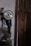 A power tool with a circular metallic blade is mounted on a wall inside a dimly lit workshop. The surroundings are rustic, showing a wooden door and several hanging objects. The surface of the wall is worn and uneven, contributing to the overall aged appearance of the setting.