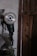 A power tool with a circular metallic blade is mounted on a wall inside a dimly lit workshop. The surroundings are rustic, showing a wooden door and several hanging objects. The surface of the wall is worn and uneven, contributing to the overall aged appearance of the setting.