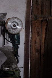 A power tool with a circular metallic blade is mounted on a wall inside a dimly lit workshop. The surroundings are rustic, showing a wooden door and several hanging objects. The surface of the wall is worn and uneven, contributing to the overall aged appearance of the setting.