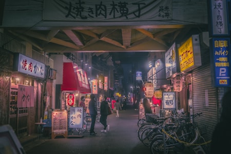 A narrow street lined with small, brightly lit shops and restaurants. Neon signs and paper lanterns hang overhead, casting a warm glow on the street. People are walking and standing near the entrances, creating a bustling, lively atmosphere. Several bicycles are parked along the sidewalk, adding to the urban setting.