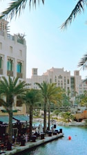 A scenic urban waterfront featuring a row of palm trees lining a canal with clear blue water. Modern Middle Eastern architecture is showcased in the buildings, with intricate designs and beige facades. Seated people are visible at outdoor cafes, adding a leisurely vibe.